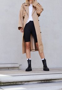 Beige Trenchcoat mit großen Knöpfen, getragen über einem weißen Oberteil und schwarzen Radshorts. Schwarze Stiefeletten mit dicker Sohle runden das Outfit ab.