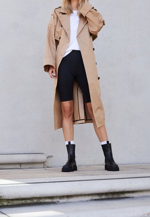 Beige trench coat with large buttons, worn over a white top and black bike shorts. Black ankle boots with a chunky sole complete the look.