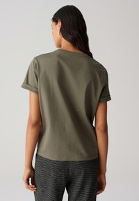 T-shirt à manches courtes vert olive avec encolure ronde et ourlets roulés, fabriqué en tissu doux ; porté avec un pantalon sombre à motifs.