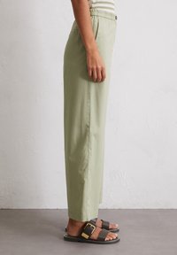 Persoon staat op een grijze vloer en draagt lichtgroene high waist-pantalons met zijzakken en donkerbruine sandalen met een grote sluiting.
