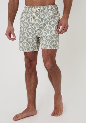 Mężczyzna w jasnoszortach kąpielowych z białymi wzorami palm i sznurkiem w pasie, stojący boso na jednolitym tle.