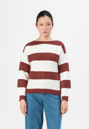 Ung kvinne med mørkt hår samlet, iført en brun og hvit stripete genser og blå jeans, stående mot en enkel hvit bakgrunn.