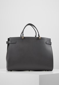 Mustat suorakulmaiset nahkalaukku kahdella kahvalla, yksinkertainen muotoilu ja kultaiset napin yksityiskohdat valkoisella pinnalla harmaata taustaa vasten.