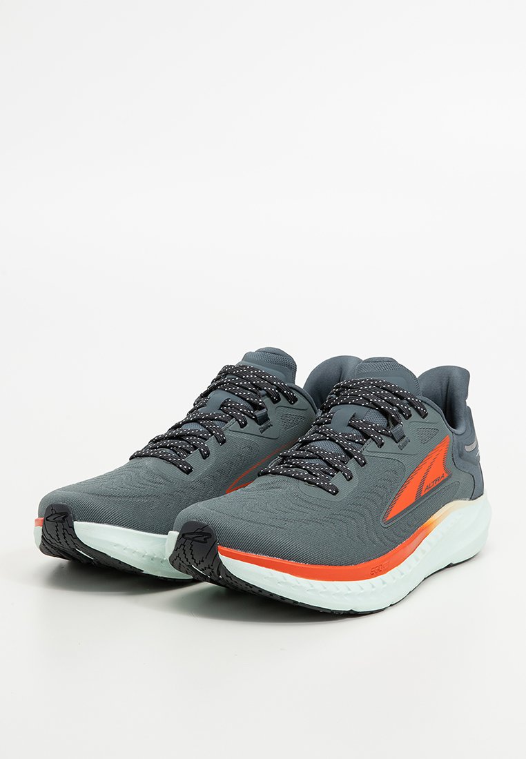 Chaussures de sport grises avec des accents orange, dessus texturé, lacets noirs et blancs à motifs, et une semelle en caoutchouc blanche.