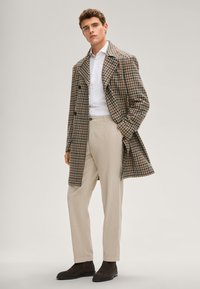 Wollmantel mit Hahnentrittmuster in Braun und Beige, mit weitem Kragen und zweireihigem Schnitt, kombiniert mit beige maßgeschneiderten Hosen und braunen Stiefeln.