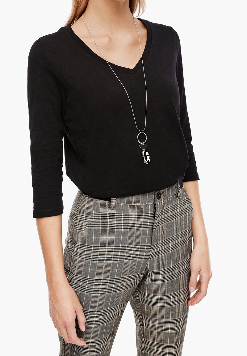 Femme portant un haut noir à col en V avec manches trois-quarts, un pantalon à carreaux taille haute, et un long collier en argent avec un pendentif noir et blanc.
