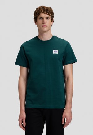 Ung mand med krøllet hår iført en mørkegrøn T-shirt med korte ærmer og en lille hvid "Lee" logo patch på brystet.