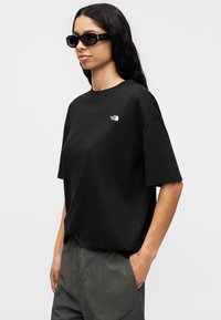 Μαύρο βαμβακερό T-shirt με κοντά μανίκια και χαλαρή εφαρμογή, με μικρό λευκό λογότυπο στο στήθος. Φοριέται με γκρι παντελόνια.