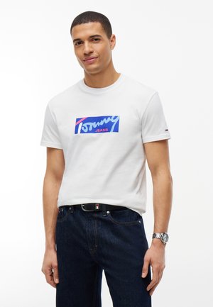 Uomo che indossa una t-shirt bianca a maniche corte con il logo Tommy Jeans blu e rosa, abbinata a jeans blu scuro e un orologio da polso argentato.