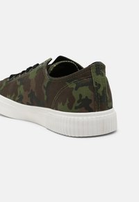 Sneaker en toile à motif camouflage avec un bout arrondi, semelle en caoutchouc blanche et lacets noirs. Présente des couleurs vert, marron et noir dans le design.