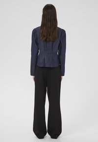 Giacca peplum in denim con maniche a sbuffo, colore blu scuro e vita aderente, abbinata a pantaloni neri larghi. Vista posteriore mostrata.