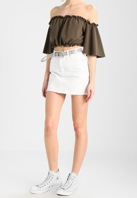 Olivově zelený crop top s odkrytými rameny a volánovým lemem, spárovaný s bílou džínovou mini sukní a bílými vysokými teniskami.