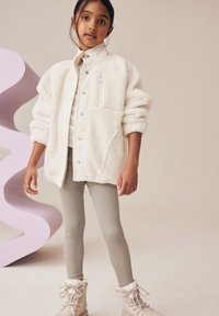 Veste en polaire blanche duveteuse, leggings beiges à côtes et bottes beiges doublées de fourrure. Textures douces et couleurs neutres. Détails de bouton simples sur la veste.