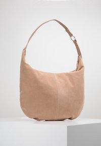 Semišová taška přes rameno v světlé béžové barvě. Měkká textura, zaoblený tvar s zakřiveným páskem a stříbrným kováním, vybavena zipovým uzávěrem.