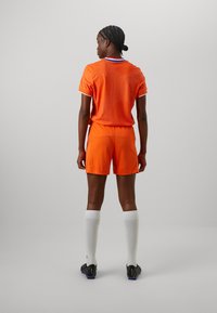Oranžový futbalový dres s mriežkovou textúrou a okrúhlym výstrihom; zodpovedajúce oranžové šortky. Biely vysoký (do kolien) ponožky a čierne futbalové kopačky dopĺňajú outfit.