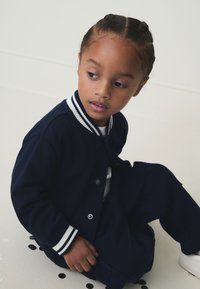 Temno modra jopica z belimi in temno modrimi črtastimi manšetami. Otrok sedi, oblečen v usklajene temno modre hlače, na jopici so vidni okrogli gumbi.