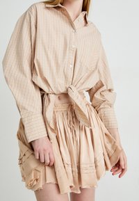 Beige plaid shirt with a tied front, paired with a light peach ruffled skirt featuring a drawstring waist and raw hem details.