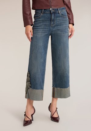 Frau trägt cropped weite blaue Jeans mit besticktem Detail und aufgerollten Säumen, kombiniert mit burgunderroten High Heels.