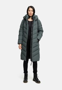 Parka lunga imbottita di colore verde scuro con cappuccio, con chiusura zip e motivi a rombi. Abbinata a pantaloni neri e stivali.