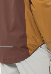 Veste colorée en marron et moutarde avec un ourlet élastique, dotée d'accents gris réfléchissants, d'une texture lisse et d'un matériau léger.