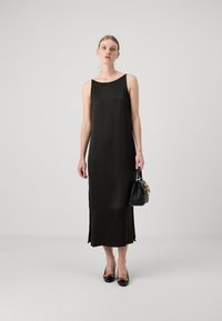 Rochie neagră, fără mâneci, din satin, cu o croială lejeră, slituri laterale și lungime până la jumătatea gambei, asortată cu o poșetă mică neagră și pantofi.