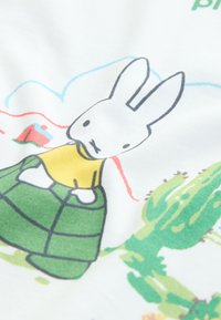 Medvilninis audinys, kuriame pavaizduotas animacinis zuikutis su geltona palaidine ir žalia raštuota suknele, apsuptas spalvingų dykumos elementų.