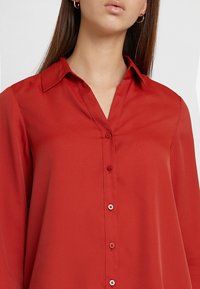 Blouse rouge à manches longues en tissu lisse. Dotée d'un col classique et d'une patte de boutonnage avec des boutons assortis.