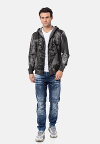 Hoodie noir zippé avec un design graphique et du texte, associé à un jean en denim bleu avec des détails cousus et des baskets grises.