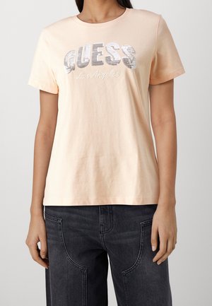 Kvinde iført en lyse fersken kortærmet t-shirt med "GUESS Los Angeles" i sølv palietter og sorte jeans.