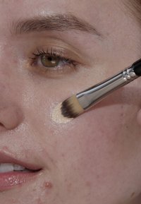 Fundamentborstel die een romige beige foundation aanbrengt op gladde, glanzende huid met kleine oneffenheden, waardoor de huidskleur wordt verbeterd.