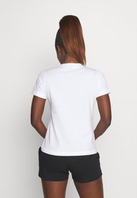 Una persona con cabello largo y castaño está de espaldas, vistiendo una camiseta blanca y pantalones cortos negros frente a un fondo sencillo.