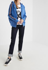 Sudadera azul con cremallera y mangas a rayas blancas y rojas, combinada con jeans de mezclilla oscura y zapatillas negras con suelas blancas.