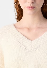 Suéter de color crema de tejido de punto con escote en V profundo. La tela parece suave y texturizada, con detalles acanalados a lo largo del escote.