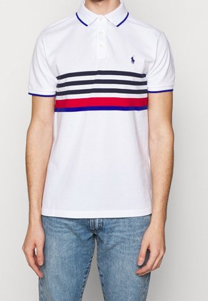 Mężczyzna w białej koszulce polo z poziomymi paskami w kolorze czarnym, czerwonym i niebieskim na wysokości klatki piersiowej, w połączeniu z jasnoniebieskimi dżinsami.