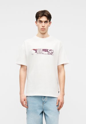 Νέος άνδρας που στέκεται με το πρόσωπο προς τα εμπρός, φοράει λευκό μπλουζάκι με αφηρημένο μοτίβο από μωβ και κόκκινα ορθογώνια και ανοιχτόχρωμο τζιν.