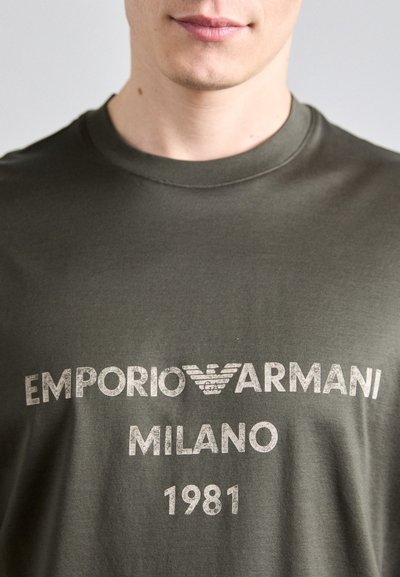 Olive rohelise puuvillase T-särgiga, millel on ümar kaelus, pleekinud valge trükitekst "EMPORIO ARMANI MILANO 1981" ja logo aktsendid.