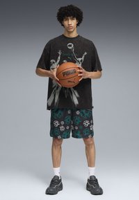 Černé grafické tričko s designem basketbalu, doplněné černými a teal vzorovanými šortkami. Model drží basketbal a má na sobě šedé tenisky.