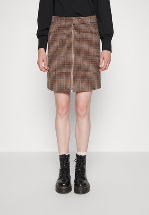 Femme portant une jupe marron à motif pied-de-poule avec fermeture éclair à l’avant, un haut noir à manches longues, des chaussettes blanches et des bottines noires à lacets, debout sur fond blanc.