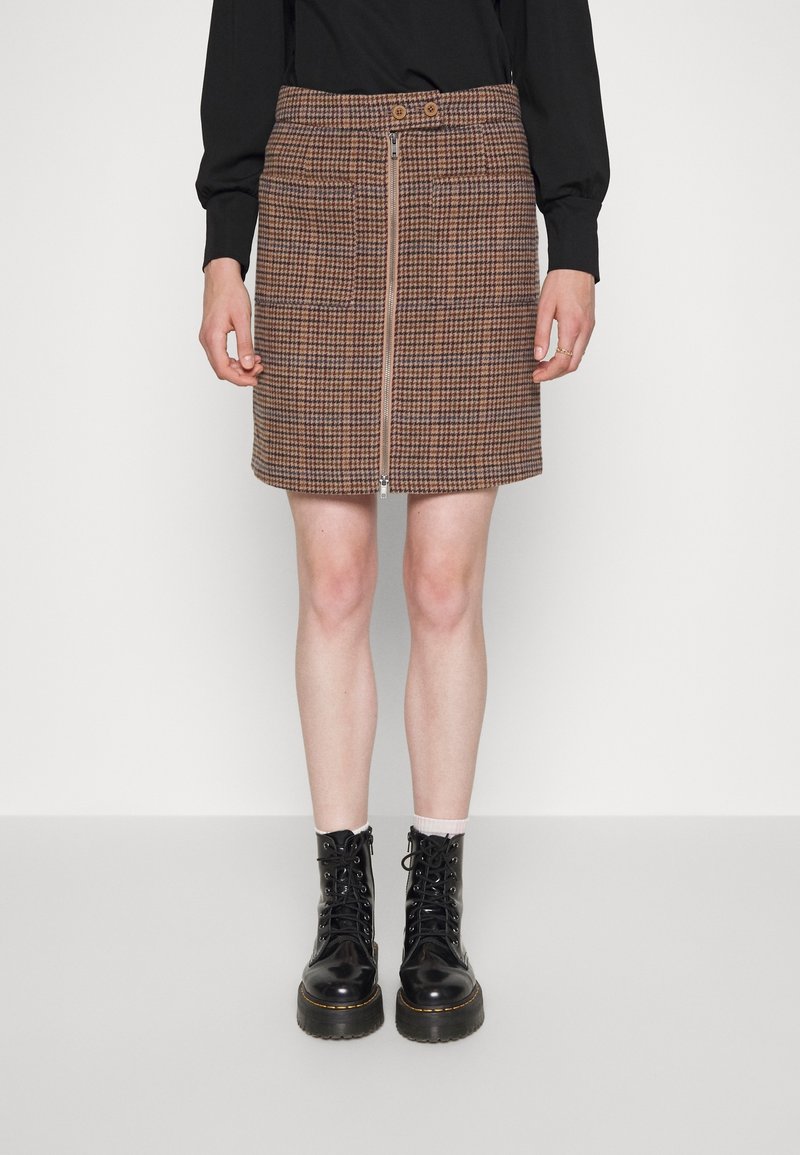 Femme portant une jupe marron à motif pied-de-poule avec fermeture éclair à l’avant, un haut noir à manches longues, des chaussettes blanches et des bottines noires à lacets, debout sur fond blanc.