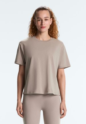 Jonge vrouw met krullend haar draagt een loszittend beige T-shirt met korte mouwen en bijpassende strakke legging, staand tegen een effen lichte achtergrond.