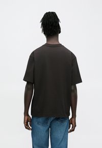 Rövid dredlokkal rendelkező személy fekete, nagy méretű pólót és kék farmert viselve, háttal áll, egyszínű háttér előtt.