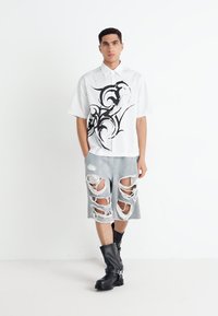 Biała koszula z krótkim rękawem w czarny wzór w spirale, zestawiona z szarymi spodniami krótkimi w stylu distressed z dużymi wycięciami oraz czarnymi kozakami do kolan.