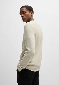 Maglione beige a maglia con un motivo testurizzato, collo a giro, e polsini a coste, abbinato a pantaloni neri. La modella è in piedi leggermente inclinata.