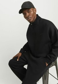 Veste zippée noire et pantalon en tissu doux, avec un col haut et des poches latérales, accompagnés d'une casquette noire assortie.