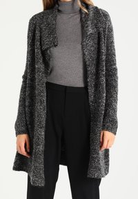 Langer, offen getragener Cardigan aus strukturiertem schwarz-grauem Strick, mit weiten Ärmeln und lockerer Passform, über einem grauen Rollkragenpullover getragen.