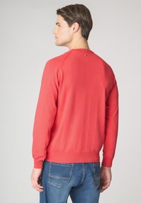 Pullover rosso a maglia con maniche lunghe, scollatura rotonda e orlo a coste. Dettaglio del logo discreto sul retro del collo. Indossato con jeans in denim blu.