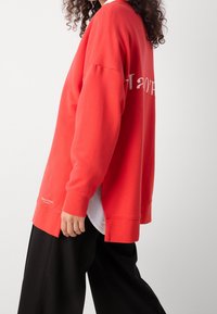 Röd oversized sweatshirt med långa ärmar, ribbade muddarna och sidofickor. Vit text på baksidan och en logotyp vid fållen. Buren över en vit topp.