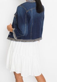 Jeansová bunda v tmavě modré barvě s roztrhaným lemem a zlatým prošitím, nošená přes bílé pruhované šaty s volány. Bunda má přední kapsy a manžety.