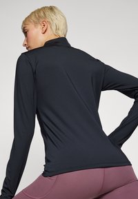Top nero a maniche lunghe in tessuto elasticizzato, con colletto alto e spalle seamless, abbinato a leggings rosa scuro.