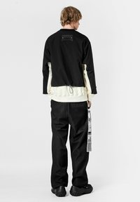 Chaqueta negra y crema con un corte holgado, que presenta un cuello alto, dobladillo elástico y un bolsillo trasero. Se lleva con pantalones negros texturizados y zapatos negros.